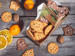A variety of cookies in a paper bag, orange chips and tangerines lie on a wooden table.