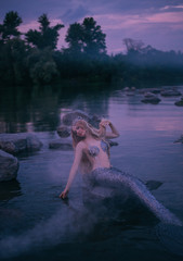 blonde siren lowers her hand, long delightful silver tail into the water, creates fog with her magic, attractive sea magician with tiara resting on stone in the middle of a quiet forest in the river