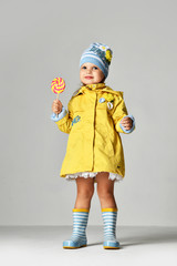 Young pretty toddler girl kid with big sweet lollypop candy in yellow jacket and rubber boots 