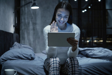 Photo of young woman sitting on bed with tablet communicating via video chat © Damir Khabirov