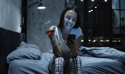 Young beautiful woman having late night video call, using her smartphone, sitting on bed, drinking...