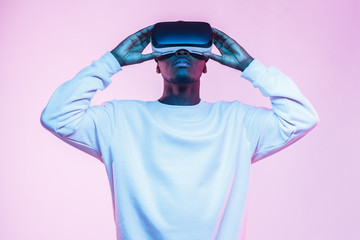Neon portrait of young african man holding virtual headset with both hands, isolated on pink...