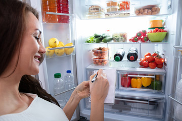 Happy Woman Writing On Spiral Notepad Near Refrigerator