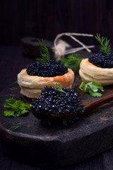 Black caviar in a wooden spoon and puff pastry tartlets on a dark board - obrazy, fototapety, plakaty