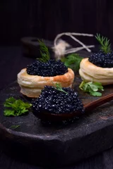 Gordijnen Black caviar in a wooden spoon and puff pastry tartlets on a dark board © kcuxen