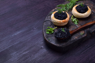 Black caviar in a wooden spoon and puff pastry tartlets on a dark board © kcuxen