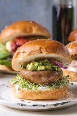 Homemade vegan vegetarian burger in wheat bun with portobello mushroom, avocado salsa and sprouts on white ceramic plate over white marble table. Close, up