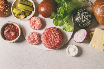 Fototapeta na wymiar Ingredients for cooking homemade hamburgers. Meat beef burger, cheese, ketchup sauce, tomato, black and white buns, salad, pickled cucumbers over grey background. Top view with space.