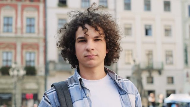 Portrait of handsome young curly man, tourist or student, millennial in hipster outfit, looking around, outdoor. Man smiling and looking at camera