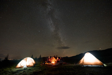 Fototapeta na wymiar Two tourist tents on mountain valley and company of happy tourists, men and women with guitar by burning campfire on background of dark starry sky with bright Milky Way constellation.