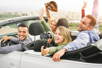 Happy friends taking photos with selfie stick camera in convertible car in vacation - Young people...