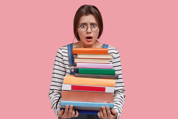 Photo of indignant young lady keeps jaw dropped from negative emotions, wears round spectacles, holds heavy pile of books, returns from library, prepares for final examination or test at college