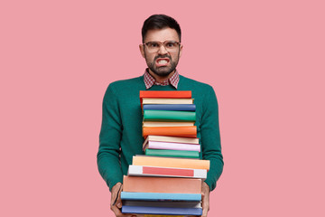 Isolated shot of irritated young man clenches teeth from anger, holds huge pile of books, wears spectacles and green sweater, does homework, prepares for lectures, stands against pink background.