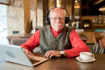 Fototapeta na wymiar Content handsome modern elderly man in headphones wearing stylish waistcoat and red turtleneck sitting at table and looking at camera while resting in cafe