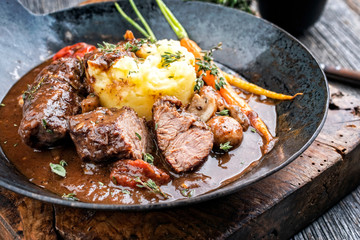 Traditionelle geschmorte Schweinebäckchen in brauner Rotwein Sauce mit Pilzen und Kartoffel Püree als closeup in einer schmiedeeisernen Pfanne