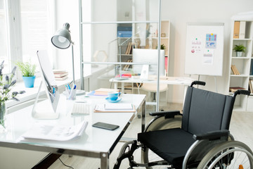 Modern office workplace with documents and computer monitor and wheelchair at the table