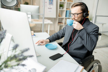 Young businessman in eyeglasses sitting in wheelchair, being disabled, listening to music on smartphone while drinking coffee and working on computer