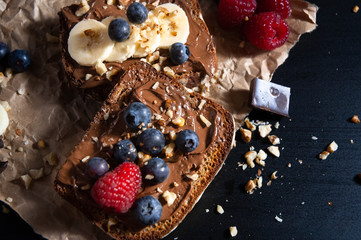 Two delicious isolated chocolate bread toast with chocolate spread, fresh banana, bluberry, rasberry and nuts toppings on rustic paper with black background