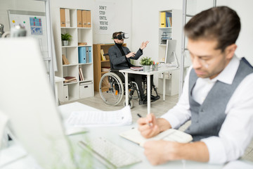 Young disabled businessman sitting in wheelchair and using virtual reality glasses while his colleague doing paperwork at the table