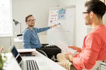 Young businessman in eyeglasses sitting in wheelchair pointing at whiteboard with graphs and talking to his partner during a presentation