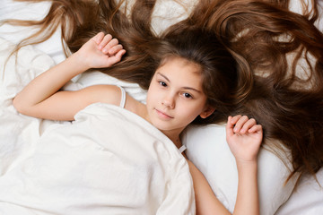 Obraz na płótnie Canvas Playful pretty girl with a long natural dark hair lies on bed covered with white soft blanket. Adorable little child spread her beautiful hair on the confortable pillow on bed
