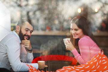 Romantic young couple in love in the restaurant with a cup of coffee talking happily together, view through a window with reflections on the glass