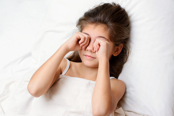 Fototapeta premium Sleepy pretty little child rubs her eyes in the morning. Lovely small child with long dark natural hair lies on comfortable bed rubbing eyes and yawning