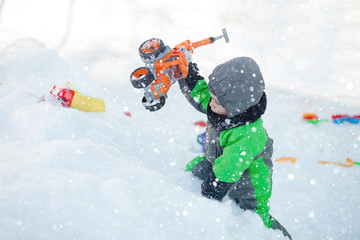 Portrait of cute little toddler sitting on snow and playing with his yellow tractor toy in the park. Child playing outdoors. Happy boy with construction toy. Lifestyle concept