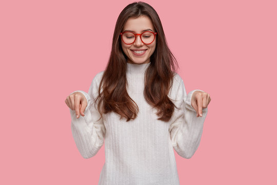 Pretty young woman with tender smile points down with both index fingers, demonstrates new shoes she bought, wears white clothes, isolated over pink background. Design and advertising concept