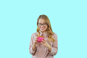 A young girl in glasses and a pink blouse holds a gift in a red box with a large bow on it and smiles with anticipation with white teeth on a blue isolated background