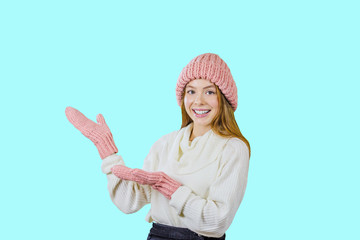 Attractive and young girl in a hat and mittens with a smile looks to the side and points with her hand on an isolated blue background