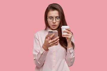 Photo of surprised dark skinned woman looks with stunned expression at screen of cell phone, has problem, recieved unpleasant news, holds disposable cup of coffee, isolated over pink background