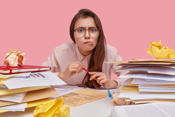 Puzzled dark haired lady purses lips, surrounded by heaps of documents, attends refresher course, wears optical glasses, isolated over pink background, works indoors. Employee difficulties concept