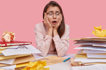 Photo of frustrated Caucasian woman with dark hair keeps hands on face, looks with puzzlement at pile of business documents, has no experience how to prepare contract, wears glasses, looks nervous