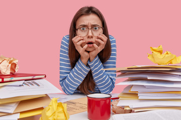 Nervous displeased lady stares directly at camera keeps hands under chin, wears blue striped jumper, square glasses, afraids of deadline, works all day with documentation, drinks hot beverage