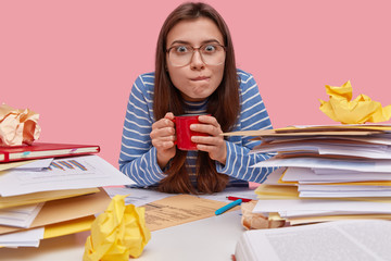 People, work, facial expressions concept. Busy brunette lady looks with suspicious attentive gaze at camera, purses lips, has coffee break, thinks about good strategy, makes report at workplace