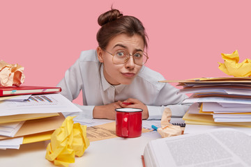 Indoor shot of attractive European student has hair knot, wears spectacles for good vision, looks scrupulously through spectacles, sits at workplace against pink background. People and working concept