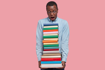 Horizontal shot of surprised dark skinned student with heap of handbooks, stares with astonishment, has to read everything for tomorrow on seminar, dressed in formal shirt and round spectacles