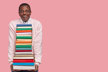Photo of attractive smiling joyful black man in casual jumper holds heap of books borrowed from...