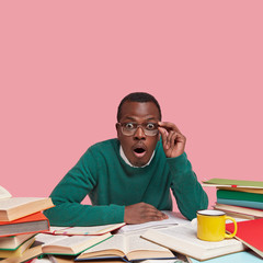 Black young male teacher looks in stupor, surprised to find out about tomorrows lecture, holds hand on rim of glasses, keeps jaw dropped from amazement, searches useful information for lesson