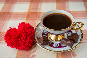 Vintage cup of coffee with heart symbol and chocolate candies on a checkered tablecloth.