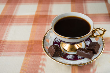 Vintage cup of coffee with chocolate candies on a checkered tablecloth.