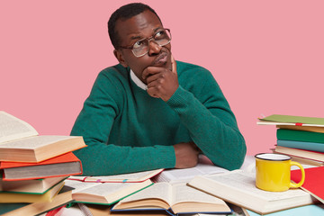 Studio shot of thoughtful black male college student focused aside, thinks on creative solution, wears casual green jumper, reads books for learning something new, poses over pink background