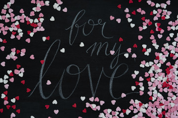 Flat lay on black chalkboard with hand lettered text message saying 'for my love' for Valentine's Day framed by small white, red and pink sugar sprinkle hearts as symbol for love and romantic feelings