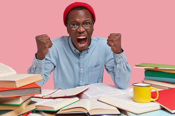 Dark skinned black man has outraged expression, clenches fists from anger, exclaims with negative emotions, wears red headgear, sits at workspace with opened books of different genres for studying