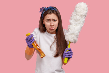 Depressed crying young woman has sorrowful expression, wears rubber gloves, carries spray and dust...