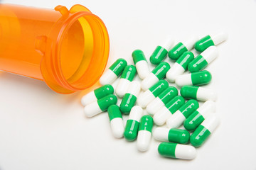 Generic-looking capsules in green and white spills out of an orange transparent bottle and empties into a plain white background in horizontal image format.