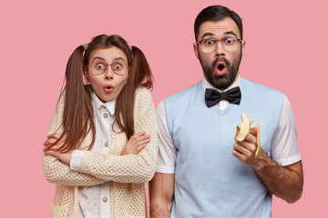 Photo of surprised female and male wonks stare with disbelief, eat delicious banana, dressed in old fashionable clothes, being puzzled by terrible news, isolated over pink background. Reaction concept