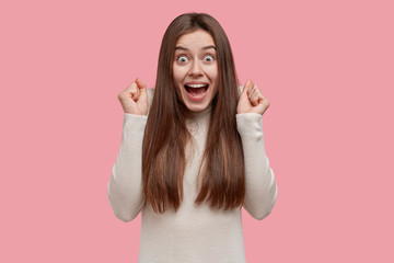 Positive European dark haired woman yells yes with success, clenches fists in triumph, celebrates victory, dressed in casual clothes, stands against pink background. People and body language concept