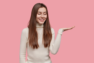 Glad brunette lady with toothy smile raises palm, pretends holding object against pink background,...